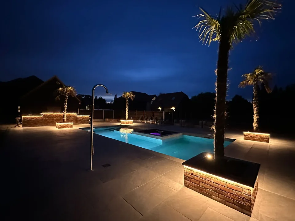 Illuminated garden planters and paved seating areas beside a swimming pool at dusk